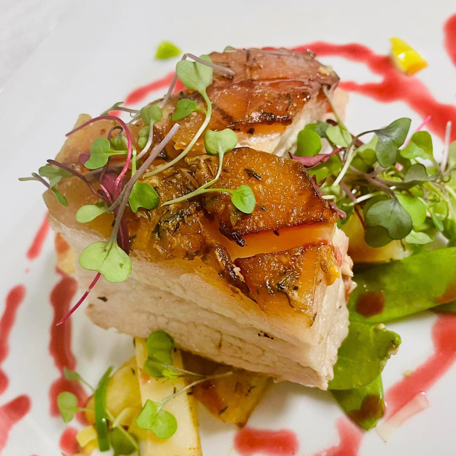 Close-up of a gourmet dish featuring crispy pork belly garnished with microgreens and accompanied by sautéed vegetables, presented on a plate with artistic sauce drizzles.
