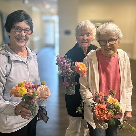 Three senior women smile brightly while holding fresh, colorful flower bouquets.