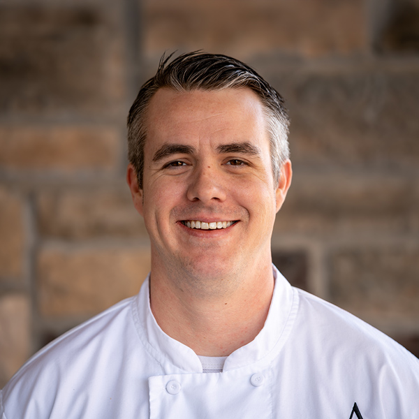 Austin Styles, Culinary Director at Pinnacle Groves Senior Living, smiling in a professional chef coat, photographed in a headshot against a softly blurred stone wall background.