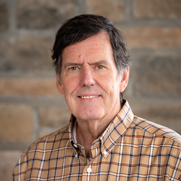 Bill Oakley, Operations Coordinator at Pinnacle Groves Senior Living, in a professional headshot wearing a tan plaid button-down shirt, standing against a softly lit stone wall background.