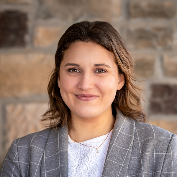 Cassidy Wagner, Assistant Executive Director at Pinnacle Groves Senior Living, smiling in a professional headshot, wearing a gray plaid blazer and white blouse, standing against a stone wall background.