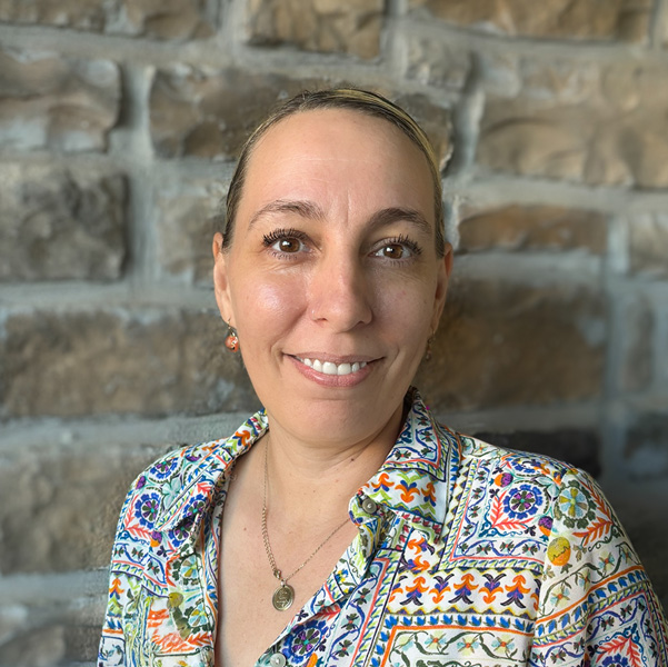 Elana Sierra, Senior Living Director at Pinnacle Groves Senior Living, wearing a colorful patterned blouse and necklace, smiling gently in front of a stone wall background.