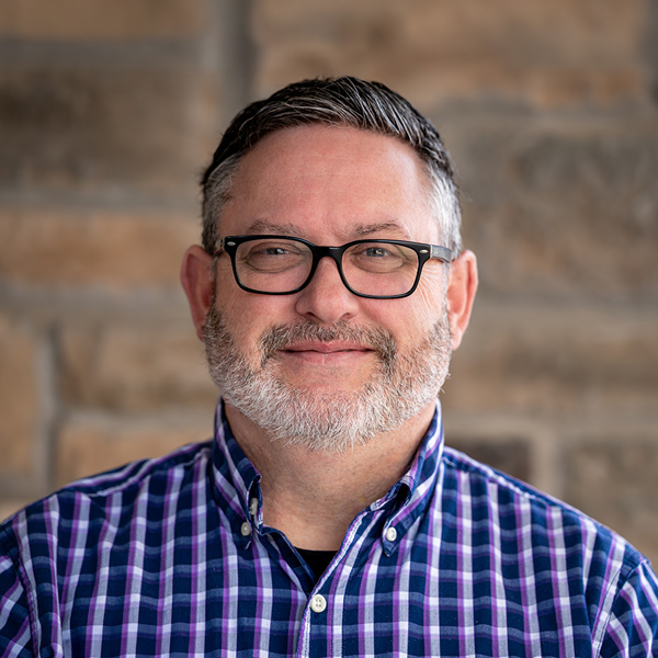 Elvin Baber, Memory Care Director at Pinnacle Groves Senior Living, wearing glasses and a purple plaid button-down, smiling in a professional headshot against a stone wall background.