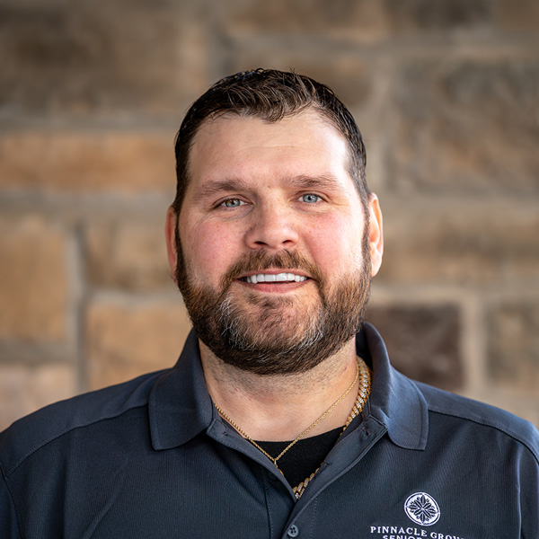 Gary Johnson, Plant Operations Director at Pinnacle Groves Senior Living, wearing a dark polo shirt, smiling in a professional headshot against a softly blurred stone wall background.