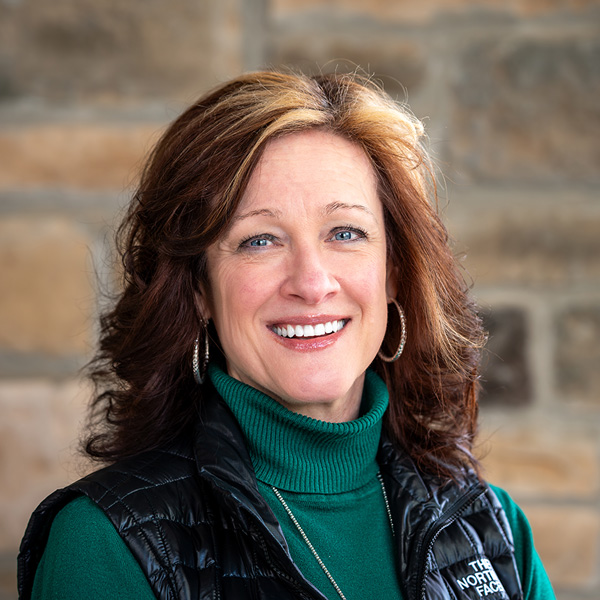 Holly Brashers, Executive Director at Pinnacle Groves Senior Living, smiling in a professional headshot, wearing a green turtleneck and black vest, standing in front of a stone wall.