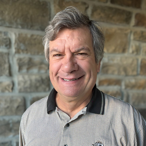 Scott Serzen, Senior Living Coordinator at Pinnacle Groves Senior Living, smiling while wearing a patterned polo shirt, standing in front of a stone wall background.