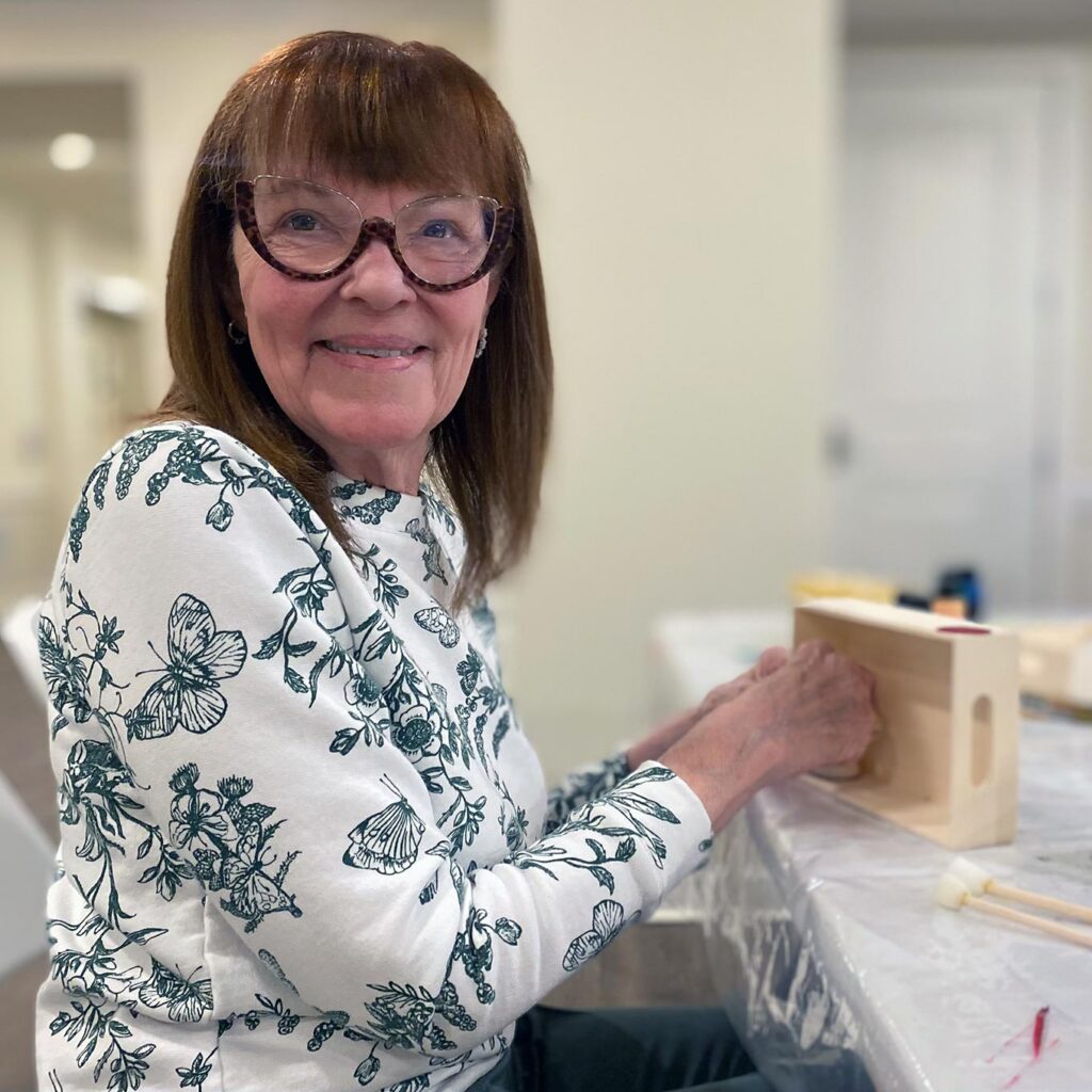 A senior woman with long, brown hair and glasses smiles while working on a crafting project.