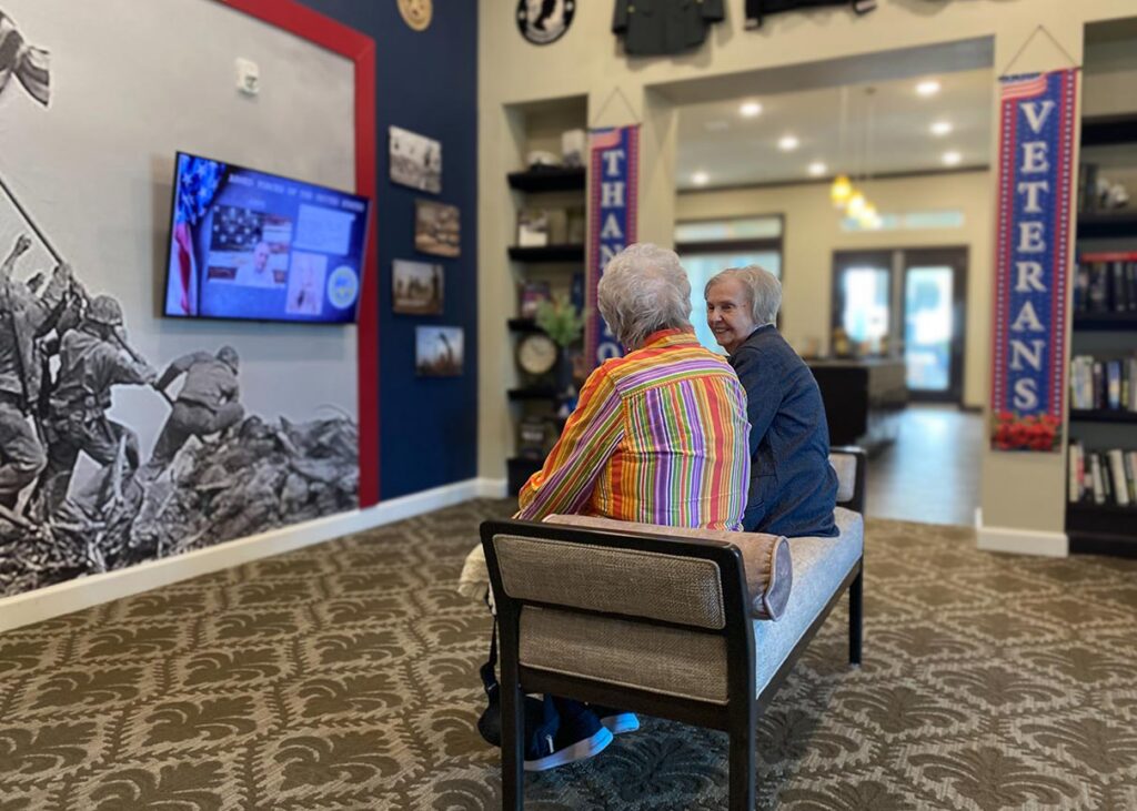 Two senior residents enjoy conversation, seated amidst a veterans display full of historical visuals and tributes to those who served.