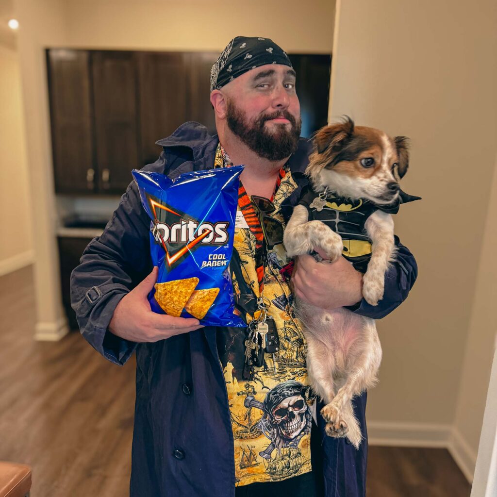 A man on the Pinnacle Groves team smiles, holding a bag of chips in one arm and a small dog dressed as a superhero in the other.
