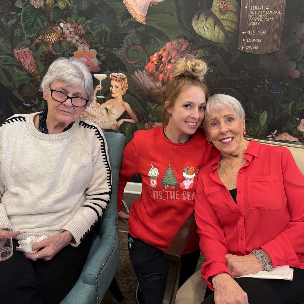 A team member and two senior women smile in front of a richly detailed backdrop at Pinnacle Groves.