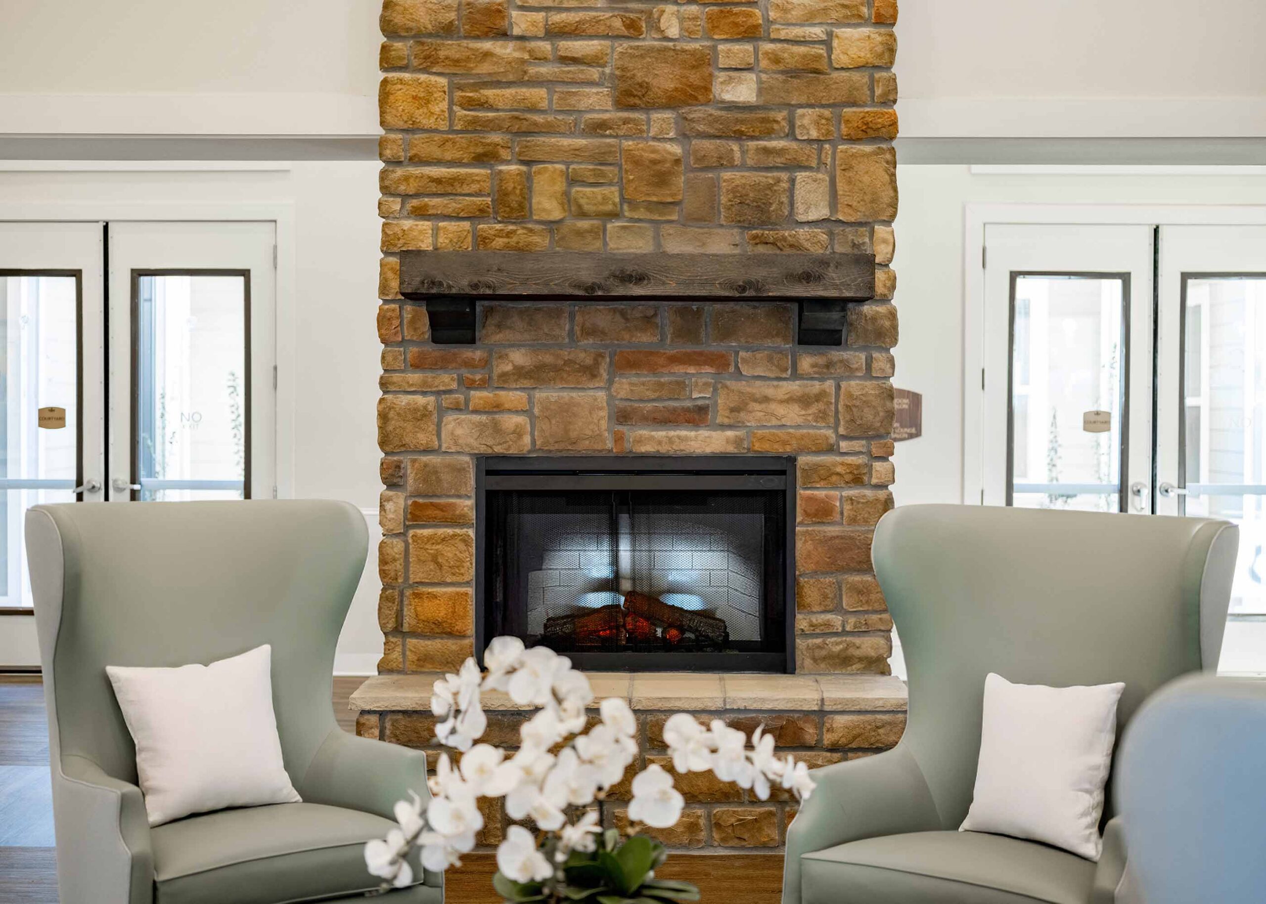 Cozy senior living seating area with stone fireplace, soft green armchairs, neutral décor, and natural light creating a warm, inviting atmosphere.
