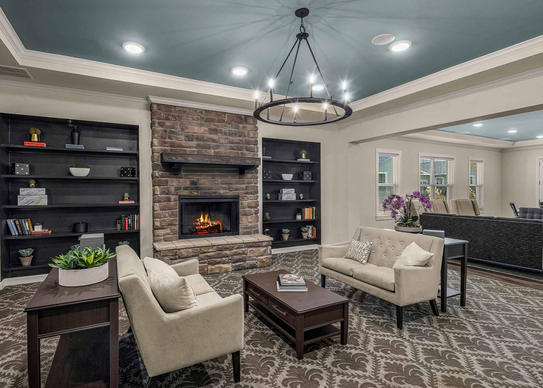 Elegant senior living lounge with stone fireplace, built-in shelving, neutral seating, patterned carpet, and soft blue ceiling with chandelier lighting.