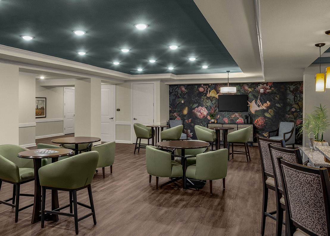 Modern senior living bistro with green upholstered chairs, wood floors, café tables, floral mural accent wall, and recessed ceiling lighting.