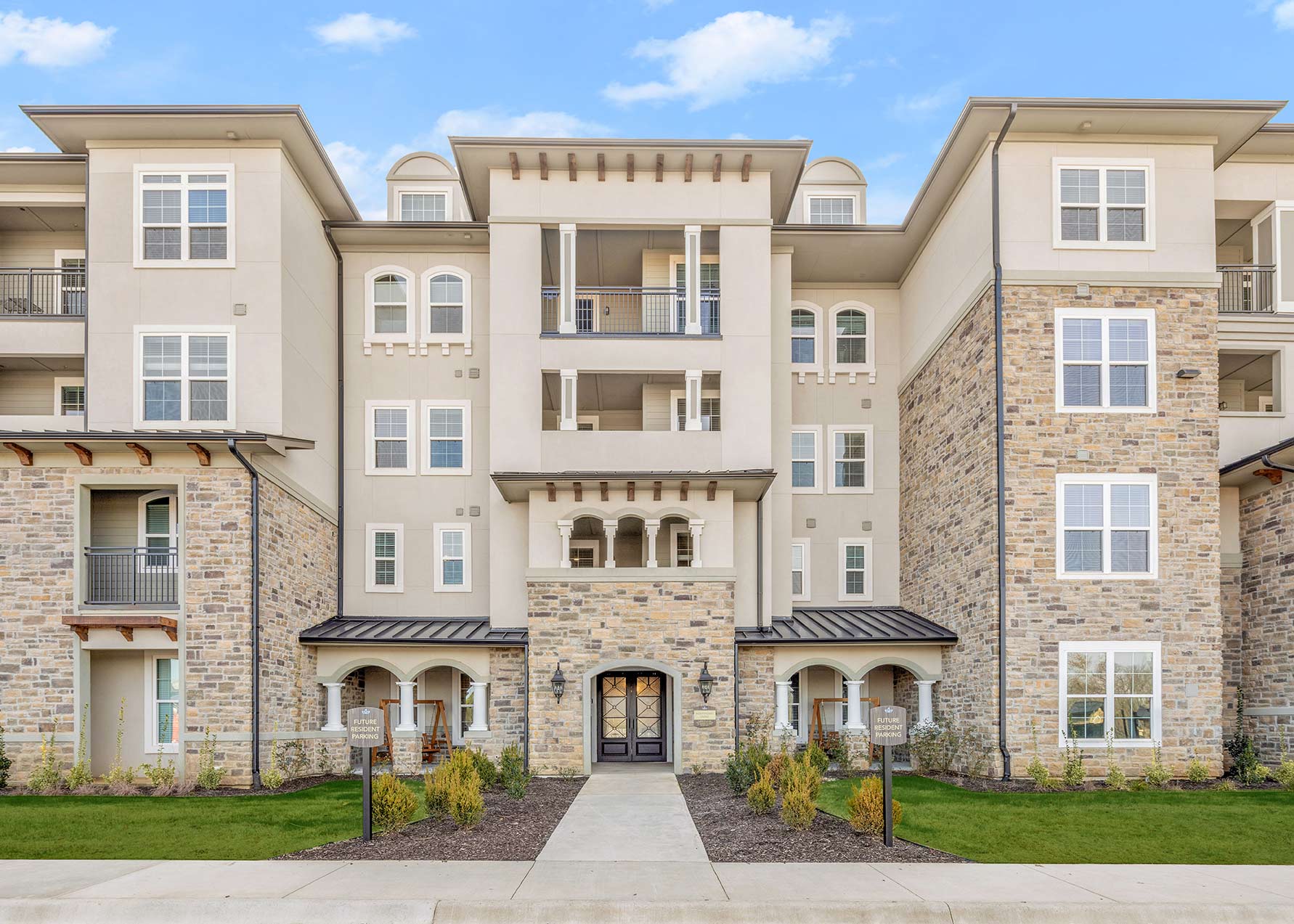 Exterior of upscale senior living community with stone and stucco facade, covered entry, balconies, and landscaped walkway leading to main entrance.