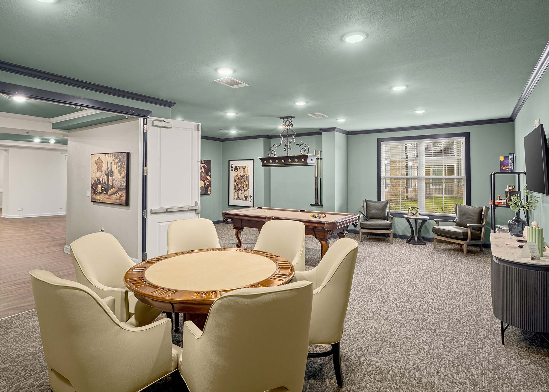 Senior living game room featuring round card table, pool table, lounge seating, soft green walls, and bright overhead lighting for social gatherings.