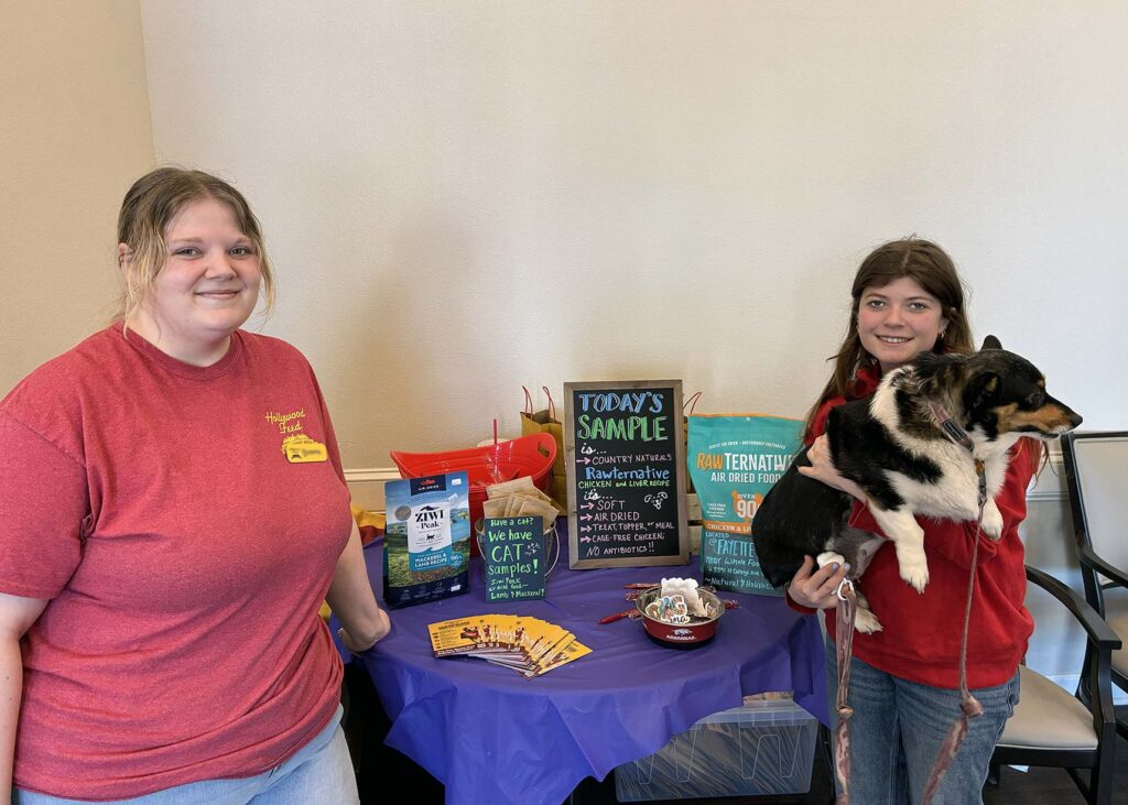 Two young women smile beside a table filled with pet product samples, one holding a tri-color Corgi.