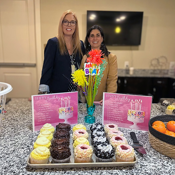 Two women smile together beside a decorated spread of gourmet cupcakes, fruit, and other treats.