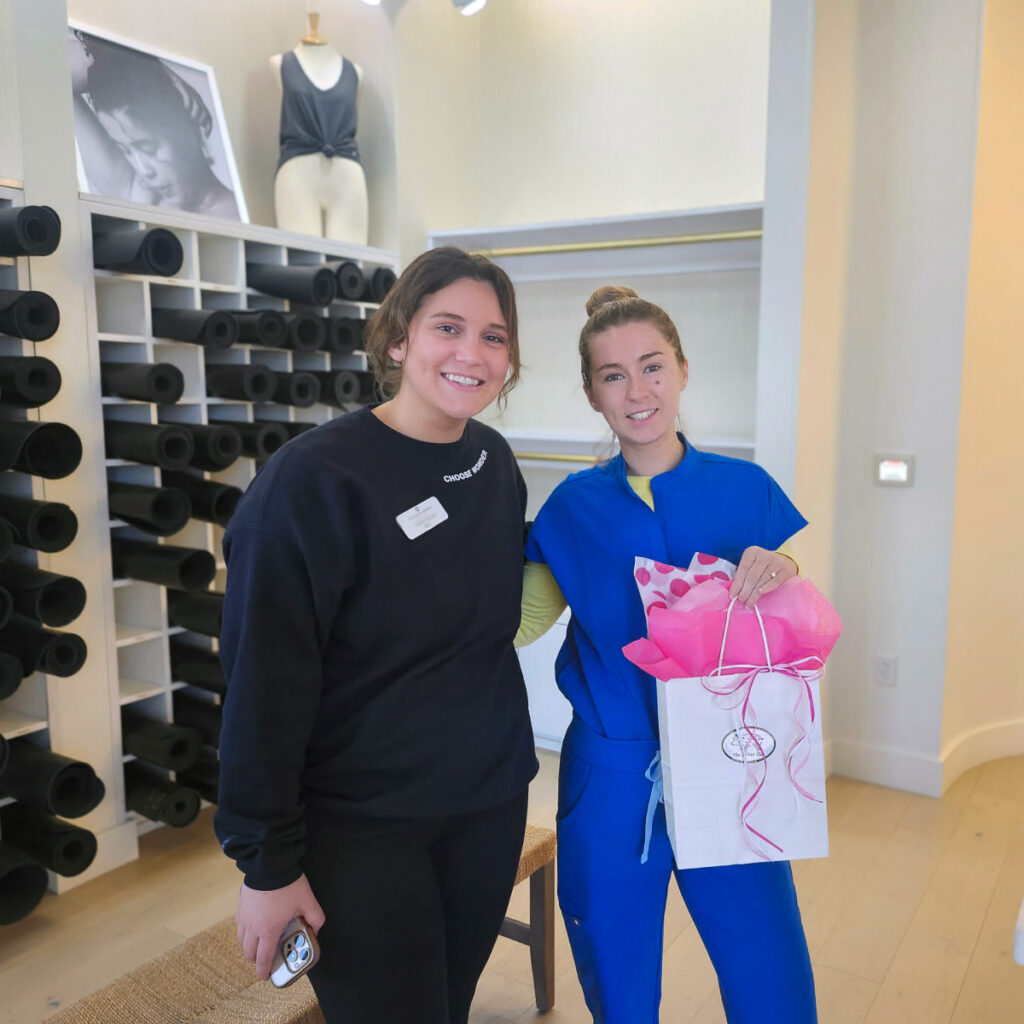 A team member from Pinnacle Groves Senior Living smiles alongside a woman from a local small business, presenting a pink gift bag.