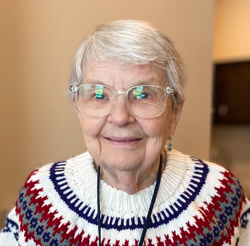 A senior woman smiles welcomingly at Pinnacle Groves Senior Living, wearing a thick patterned sweater and glasses.