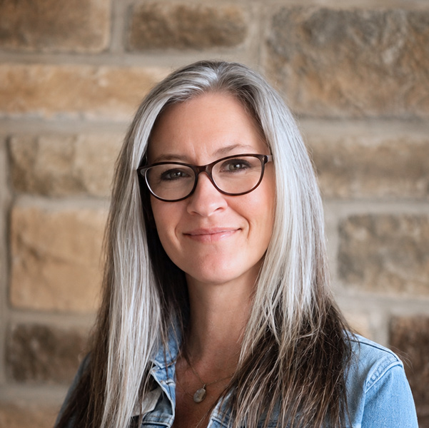 Kacy Dodge, Business Office Director at Pinnacle Groves Senior Living, looking at the camera in a professional headshot with long silver-gray hair, glasses, a denim jacket, and a tan stone wall background.